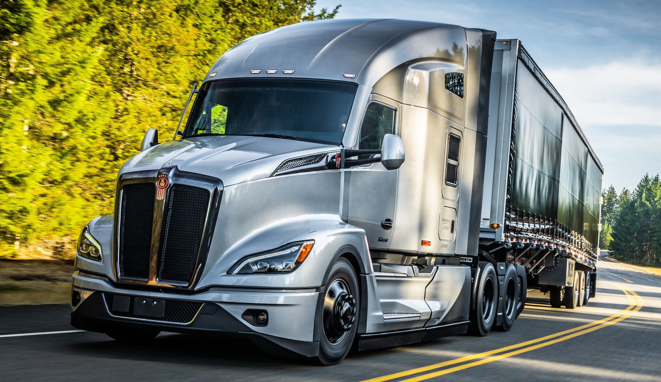 A silver Kenworth semi truck driving on a road surrounded by trees.
