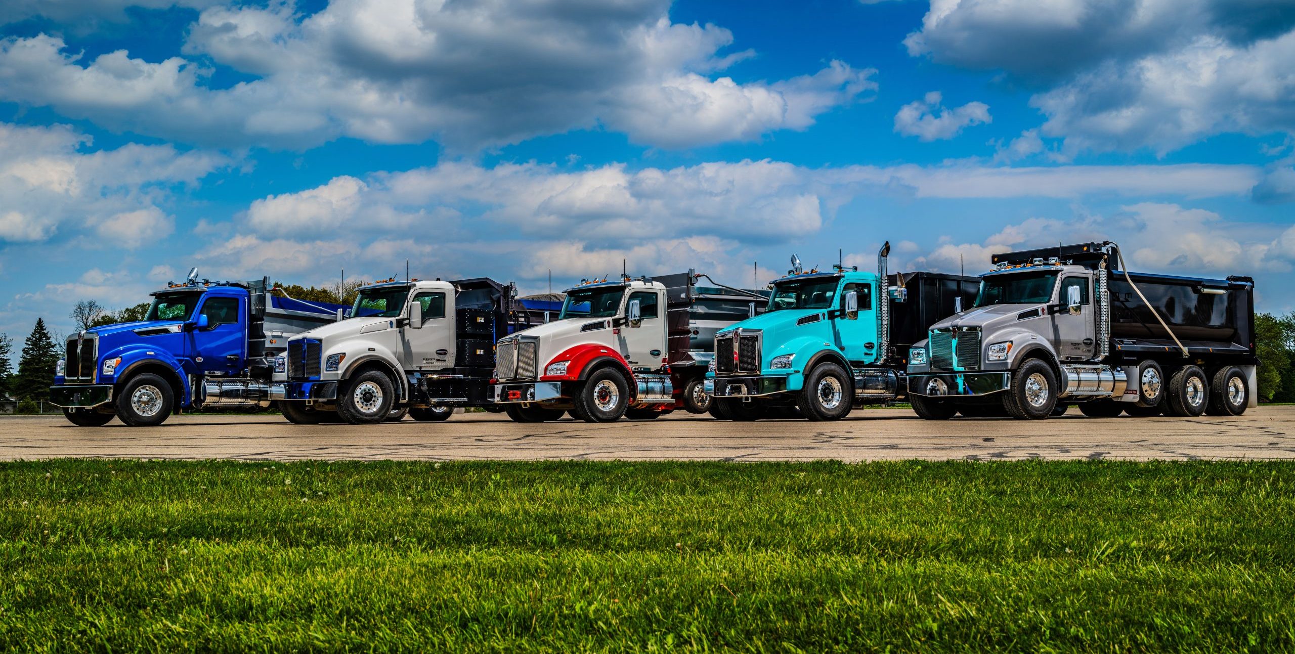 Five Kenworth Trucks lined up on grass with blue skies in the background